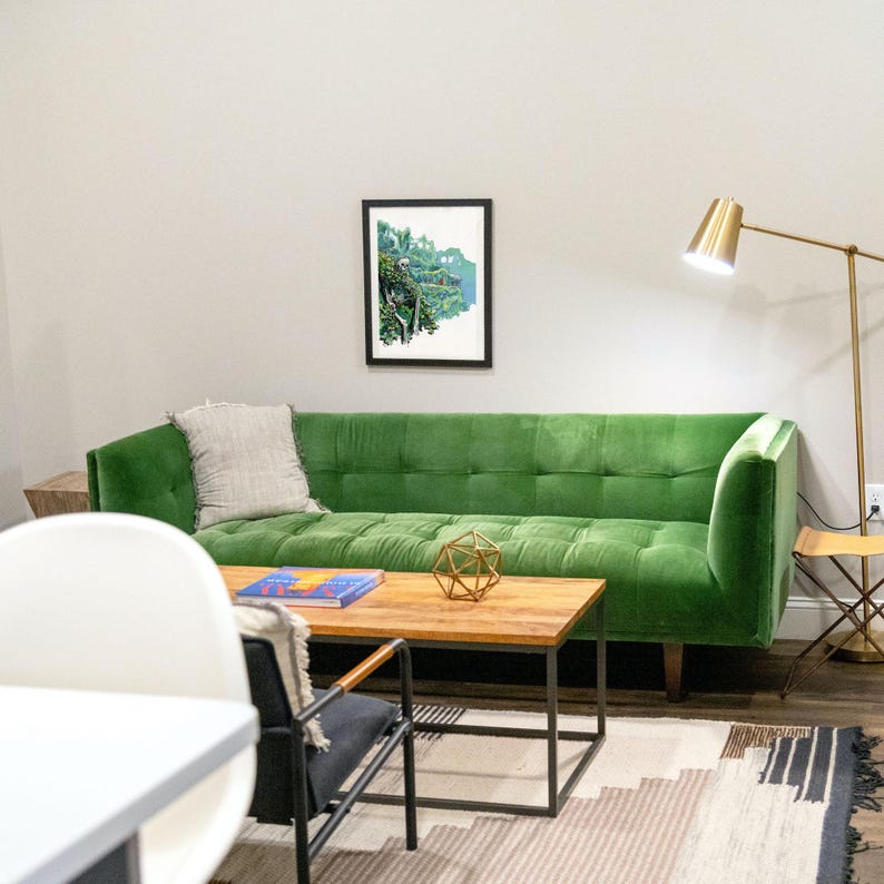 May include: A green velvet sofa with a gray throw pillow sits in a living room with a wooden coffee table and a gold floor lamp. A framed print of a landscape hangs on the wall.