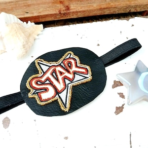 May include: Black eye patch with a gold, red, and white star design. The eye patch has a black elastic band. A seashell and a star-shaped decoration are in the background.