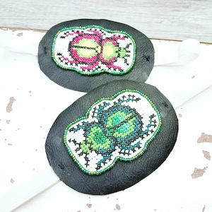 May include: Two black leather eye patches with embroidered beetle designs. One patch features a pink and green beetle, the other a green and blue beetle. Both have white elastic straps for wear.