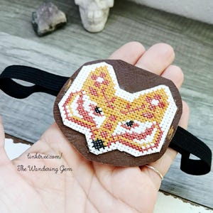 May include: A brown leather eye patch with a cross-stitched fox design in orange, red, and white. It has a black elastic strap. The text "linktr.ee/ The Wandering Gem" is visible on the patch.