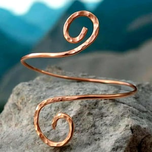 May include: A copper-colored arm cuff with a hammered texture. The cuff features two spiral designs at each end, connected by a gently curved band. The jewelry is displayed on a gray rock with a blurred blue and green background.