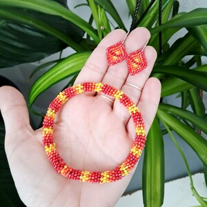 Puede incluir: Un conjunto de joyas de cuentas con una pulsera y pendientes a juego. La pulsera y los pendientes son rojos, amarillos y naranjas, con un patrón geométrico. Los pendientes tienen forma de diamante y cuelgan de ganchos plateados. La pulsera es circular.