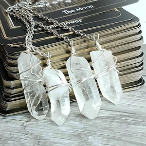 May include: Five clear quartz crystal pendants wrapped in silver wire, hanging from silver chains. The pendants are arranged in a row on a stack of tarot cards, with the card "The Moon" visible at the top.