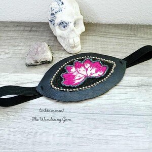 May include: Black eye patch with a pink and white floral design. The patch is embellished with silver beads and a black elastic band. A decorative skull and a crystal are in the background.