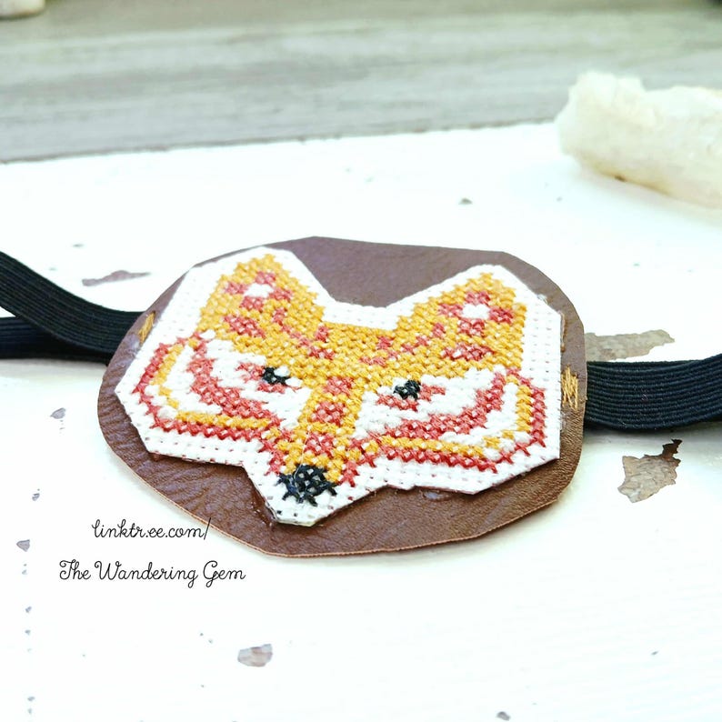 May include: A brown leather eye patch featuring a cross-stitched fox design in orange, yellow, white, and red. It has a black elastic strap. The text "linktr.ee/ The Wandering Gem" is printed on the patch.