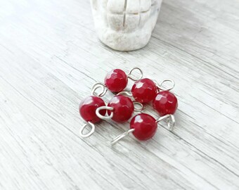 May include: Seven faceted, deep red beads with silver-colored loops are clustered together. A carved, white stone skull is in the background. The beads are on a light gray wooden surface.