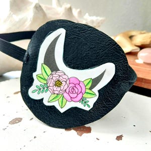 Può includere: Fascetta per occhi nera con un design floreale. La toppa presenta un design a corno di unicorno bianco e nero con fiori rosa e foglie verdi. È attaccata una fascia elastica da indossare.