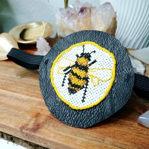May include: A round, dark gray hair tie with a stitched bee design. The bee is yellow and black, surrounded by a yellow border on a white background. The hair tie is on a wooden surface.