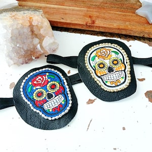 Puede incluir: Dos pulseras de cuero negro con diseños de calaveras de azúcar del Día de Muertos. Una pulsera presenta un diseño floral azul y rojo, mientras que la otra tiene un diseño floral amarillo y naranja. Ambas tienen bandas elásticas.
