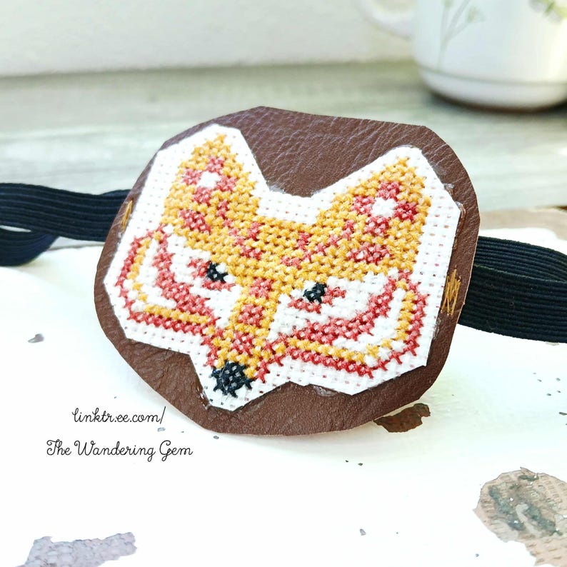 May include: A brown leather patch featuring a cross-stitched fox face in yellow, orange, red, and white. The patch is attached to a black elastic band. The text "linktr.ee.com/ The Wandering Gem" is visible.