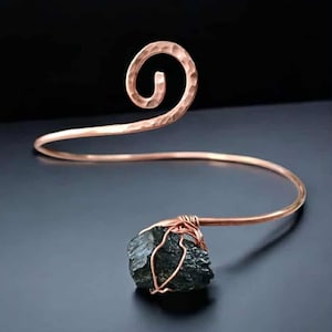 May include: A copper-colored arm cuff with a hammered texture and a spiral design. The cuff features a dark, irregularly shaped stone wrapped in copper wire. The jewelry is set against a dark background.