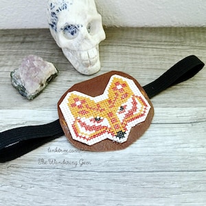 May include: A brown leather eye patch with a cross-stitched fox design in orange, red, and white. The patch is attached to a black elastic band. The text "linktr.ee/ The Wandering Gem" is visible.