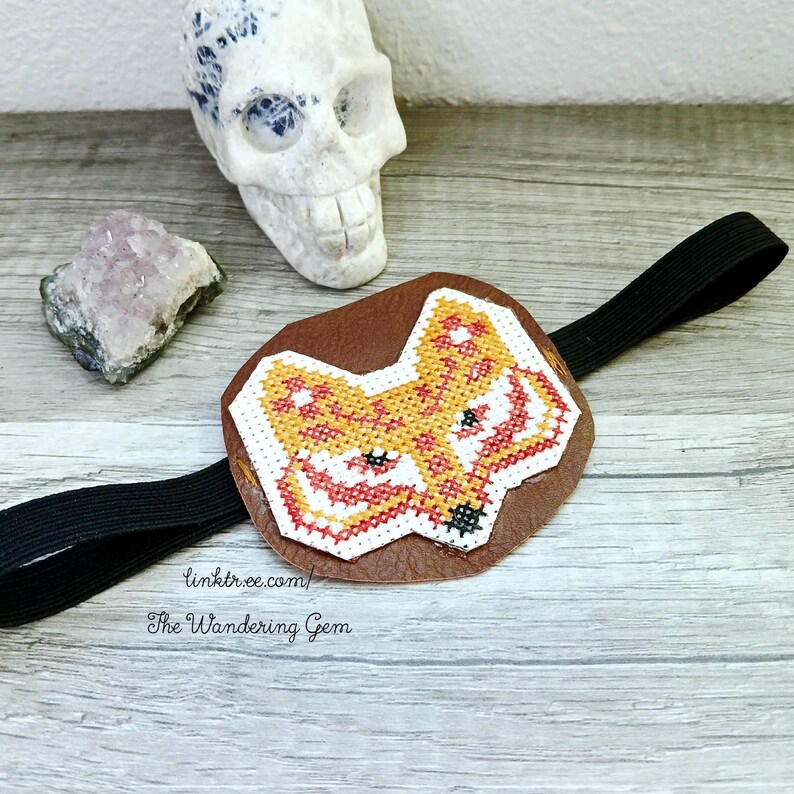 May include: A brown leather eye patch featuring a cross-stitched fox design in orange, red, and white. It is attached to a black elastic band. The text "linktr.ee/ The Wandering Gem" is visible.