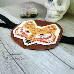 May include: A brown leather eye patch featuring a cross-stitched fox design in orange, red, and white. It has a black elastic strap. The text "linktr.ee.com/ The Wandering Gem" is printed on the patch.