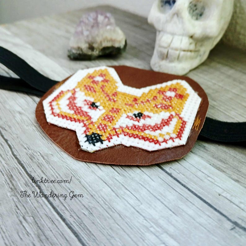May include: A brown leather eye patch featuring a cross-stitched fox design in orange, red, and white. It has a black elastic strap. The text "linktr.ee.com/ The Wandering Gem" is printed on the patch.