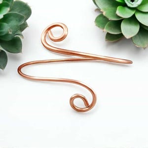 May include: A copper-colored hair accessory with a curved design. The hair pin features two spiral ends and a straight bar in the middle. The accessory is displayed on a white surface with green plants in the background.