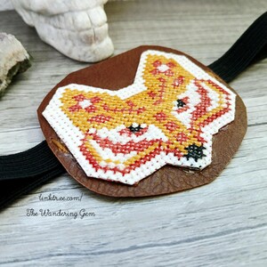 May include: A brown leather eye patch featuring a cross-stitched fox design in orange, red, and white. The fox has black eyes and a black nose. The eye patch has a black elastic band. The text "linktr.ee/ The Wandering Gem" is visible.