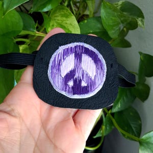 May include: Black eye patch with a purple embroidered peace sign. The patch has an elastic band for secure wear. The peace sign is in a circle and is made with purple thread. The eye patch is made of black material.