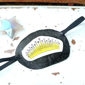 Puede incluir: Parche ocular negro con un diseño de corona amarilla y morada. El parche ocular tiene una banda elástica negra y está hecho de un material texturizado. Un objeto translúcido en forma de estrella está en el fondo.