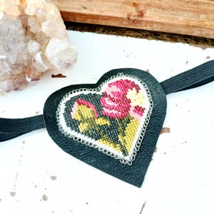 May include: A black heart-shaped eye mask with a floral cross-stitch design in pink, yellow, and green. The mask has a white lace trim and a black elastic band. The mask is approximately 7.6 cm wide.