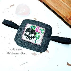 Pode incluir: Tampão de olho preto com um desenho de caveira e rosa bordada. A caveira é preta e a rosa é rosa com folhas verdes. Possui uma faixa elástica preta. O texto "linktr.ee.com/ The Wandering Gem" está impresso na parte inferior.
