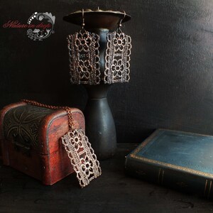 Puede incluir: Un conjunto de tres delicados pendientes rectangulares de color cobre y un collar con un colgante a juego. Los pendientes y el colgante están hechos de intrincados diseños de encaje. Las joyas se muestran en un soporte de madera y un libro antiguo.