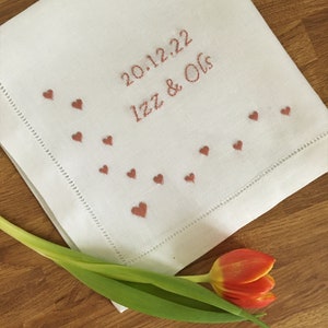 K&ouml;nnte beinhalten: Wei&szlig;es Leinentaschentuch, bestickt mit dem Datum "20.12.22" und den Namen "Izz & Ols" in rotbraunem Faden. Kleine Herzformen sind am Rand gestickt. Eine rot-gelbe Tulpe liegt auf einer Holzoberfl&auml;che.