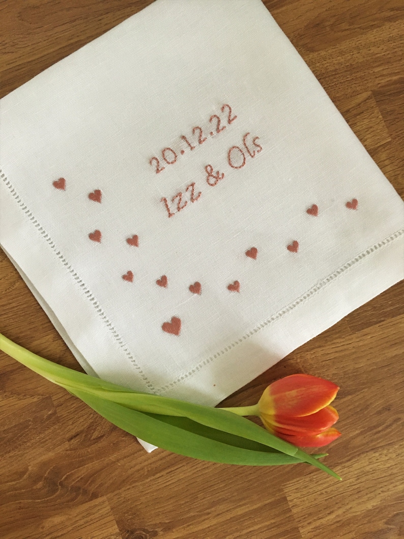 K&ouml;nnte beinhalten: Wei&szlig;es Leinentaschentuch, bestickt mit dem Datum "20.12.22" und den Namen "Izz & Ols" in rotbraunem Faden. Kleine Herzformen sind am Rand gestickt. Eine rot-gelbe Tulpe liegt auf einer Holzoberfl&auml;che.