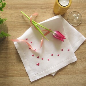 K&ouml;nnte beinhalten: Eine wei&szlig;e Leinen-Serviette mit dekorativem Rand, verziert mit kleinen roten herzf&ouml;rmigen Verzierungen. Eine rosa Tulpe und ein rosa Band liegen auf der Serviette. Ein Kerzenhalter und ein Weinglas befinden sich im Hintergrund.