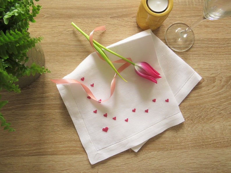 K&ouml;nnte beinhalten: Eine wei&szlig;e Leinen-Serviette mit dekorativem Rand, verziert mit kleinen roten herzf&ouml;rmigen Verzierungen. Eine rosa Tulpe und ein rosa Band liegen auf der Serviette. Ein Kerzenhalter und ein Weinglas befinden sich im Hintergrund.