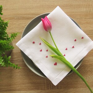 K&ouml;nnte beinhalten: Eine wei&szlig;e Leinen-Serviette mit gesticktem Rand, verziert mit kleinen roten herzf&ouml;rmigen Streuseln, liegt auf einem grauen Teller. Eine rosa Tulpe mit gr&uuml;nen Bl&auml;ttern liegt auf der Serviette. Im Hintergrund befindet sich ein Topf mit Farn.