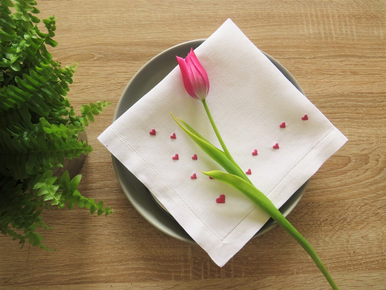 K&ouml;nnte beinhalten: Eine wei&szlig;e Leinen-Serviette mit gesticktem Rand, verziert mit kleinen roten herzf&ouml;rmigen Streuseln, liegt auf einem grauen Teller. Eine rosa Tulpe mit gr&uuml;nen Bl&auml;ttern liegt auf der Serviette. Im Hintergrund befindet sich ein Topf mit Farn.