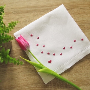 K&ouml;nnte beinhalten: Wei&szlig;es Leinen-Taschentuch mit gestickten roten Herzen. Eine rosa Tulpe und ein gr&uuml;ner Stiel liegen auf einer Holzoberfl&auml;che. Das Taschentuch hat eine dekorative Hohlsaum. Romantisches und elegantes Design.