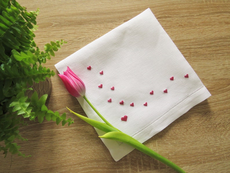 K&ouml;nnte beinhalten: Wei&szlig;es Leinen-Taschentuch mit gestickten roten Herzen. Eine rosa Tulpe und ein gr&uuml;ner Stiel liegen auf einer Holzoberfl&auml;che. Das Taschentuch hat eine dekorative Hohlsaum. Romantisches und elegantes Design.