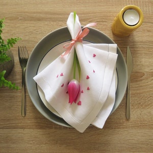 K&ouml;nnte beinhalten: Tischarrangement mit einer wei&szlig;en Leinen-Serviette, verziert mit rosa herzf&ouml;rmigen Konfetti und einer rosa Tulpe. Die Serviette ist mit einem rosa Band gebunden und auf einem grauen Teller platziert. Eine Gabel und ein Messer sind vorhanden.