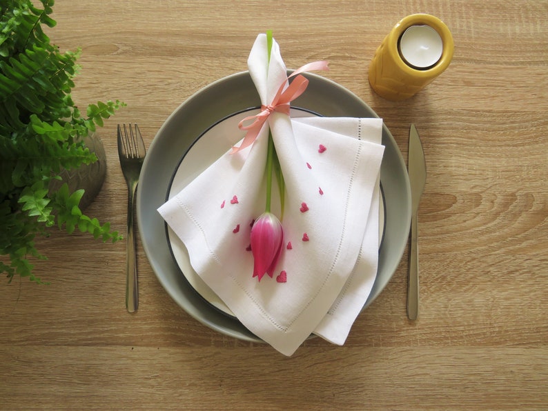 K&ouml;nnte beinhalten: Tischarrangement mit einer wei&szlig;en Leinen-Serviette, verziert mit rosa herzf&ouml;rmigen Konfetti und einer rosa Tulpe. Die Serviette ist mit einem rosa Band gebunden und auf einem grauen Teller platziert. Eine Gabel und ein Messer sind vorhanden.