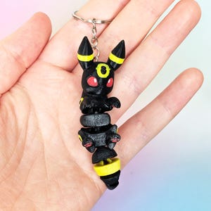 Könnte beinhalten: Ein schwarzer und gelber Umbreon-Schlüsselanhänger. Die Umbreon-Figur hat rote Augen und gelbe Akzente an Ohren und Schwanz. Der Schlüsselanhänger ist aus Kunststoff und an einem silbernen Metallring befestigt.