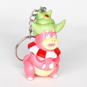 Könnte beinhalten: Ein rosa und grüner Pokemon-Schlüsselanhänger mit einer Figur mit grünem Hut und rotem Schal.