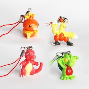 May include: Four colourful Pokemon character keychains with attached strings. The characters are a red and yellow bird, a yellow and orange bird, a red dinosaur, and a green lizard.