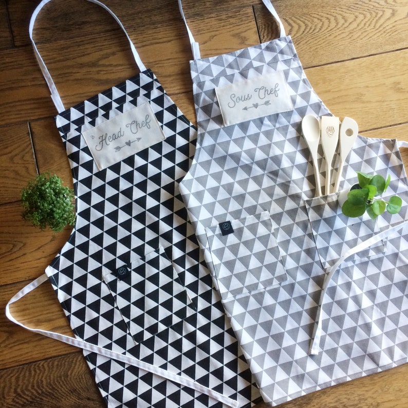 Grey and white triangle and black and white triangle pattern aprons from quality cotton with hand printed Head chef and sous chef tag and arrow with heart design, plus wooden cooking tools, all from My Home and Yours.