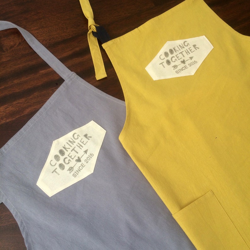 Custom linen cotton aprons premium size in mustard yellow and grey, with hand printed cooking together since tag to which you can add the date. Unique quality couple anniversary gift.