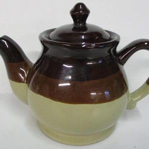 May include: A ceramic teapot with a lid, spout, and handle. The teapot features horizontal bands of dark brown, medium brown, and light beige. The handle and spout have a light beige accent. The teapot is designed for brewing tea.