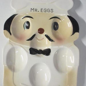 May include: A novelty ceramic egg holder in the shape of a chef, featuring the text "MR. EGGS". The egg holder is predominantly white with black details, a black bow tie, and a yellow base. The chef has a mustache and rosy cheeks.