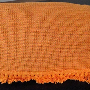 May include: Orange woven throw blanket with a fringe trim. The blanket has a textured, grid-like pattern and is folded neatly. The fringe is a matching orange colour. The blanket is made of a soft material.