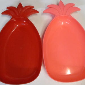 May include: Two pineapple-shaped serving dishes made of smooth plastic. One dish is a deep, vibrant red colour, while the other is a lighter, coral-pink shade. Both feature a stylised leaf design at the top and a rounded, oval-shaped basin for holding items.