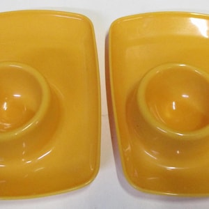 May include: Two matching rectangular egg cups with a smooth, glossy finish in a vibrant mustard yellow colour. Each piece features a central circular indentation designed to hold a single egg, integrated into a flat, wide base with rounded corners.