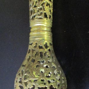 May include: An ornate brass vase with a filigree design. The vase features a bulbous base and a slender, openwork body. The base is supported by three decorative feet. The overall height is approximately 15 cm.