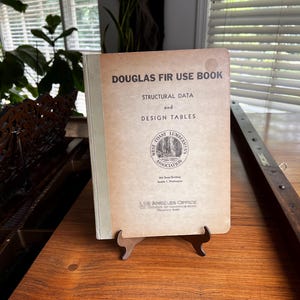 May include: A vintage "Douglas Fir Use Book" is displayed on a wooden surface. The book has a tan cover with black text, including "Structural Data and Design Tables." The West Coast Lumbermen's Association logo is visible.