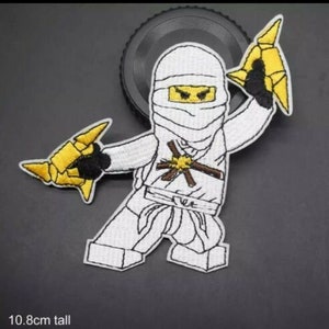 Könnte beinhalten: Gestickter Aufnäher mit einem weißen Ninja-Charakter mit gewickeltem Kopf und gelben Augen. Der Ninja hält zwei gelbe Wurfsterne. Der Aufnäher ist 10,8 cm hoch und hat einen schwarzen Rand.