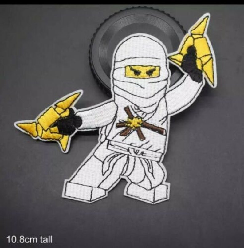 May include: Embroidered patch of a white ninja character with a wrapped head and yellow eyes. The ninja holds two yellow throwing stars. The patch is 10.8cm tall and has a black border.